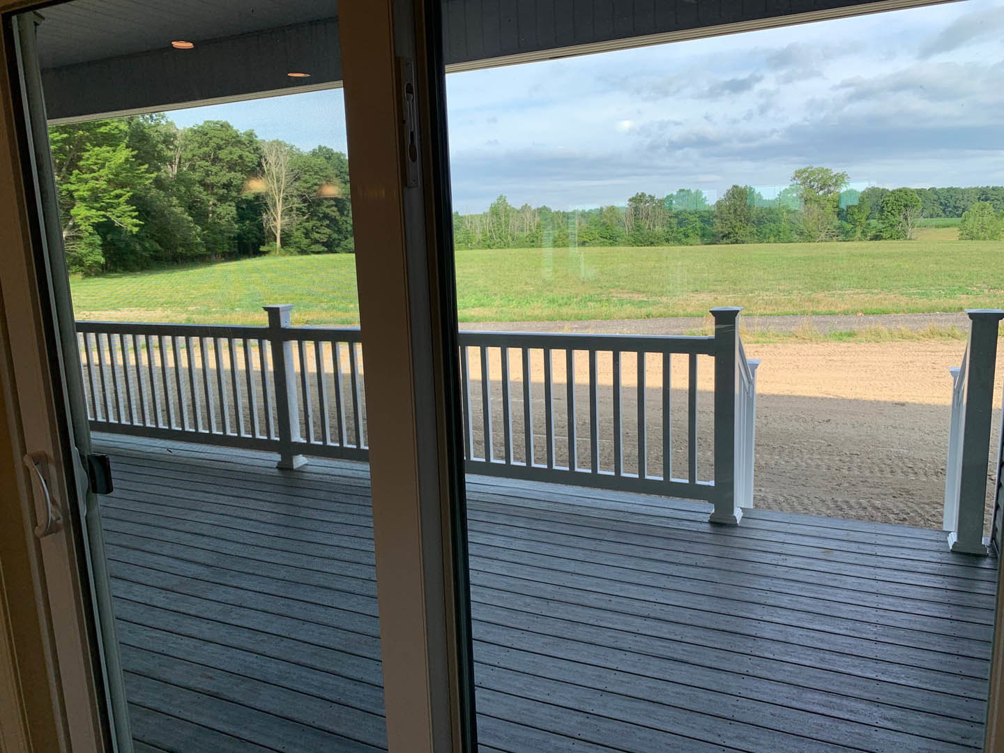 Wood deck with metal railing overlooking green grass field, sliding glass door, and blue sky visible through window
