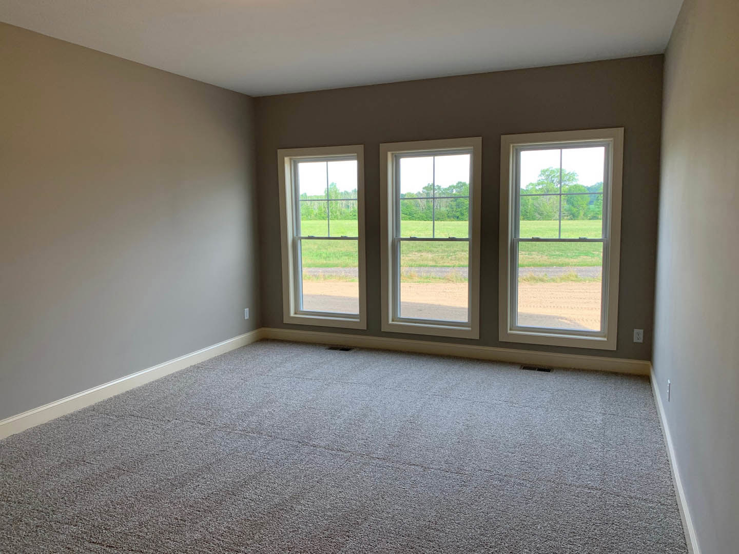 Carpeted room with three large windows overlooking grassy yard and trees, white walls, floor vent visible near window.