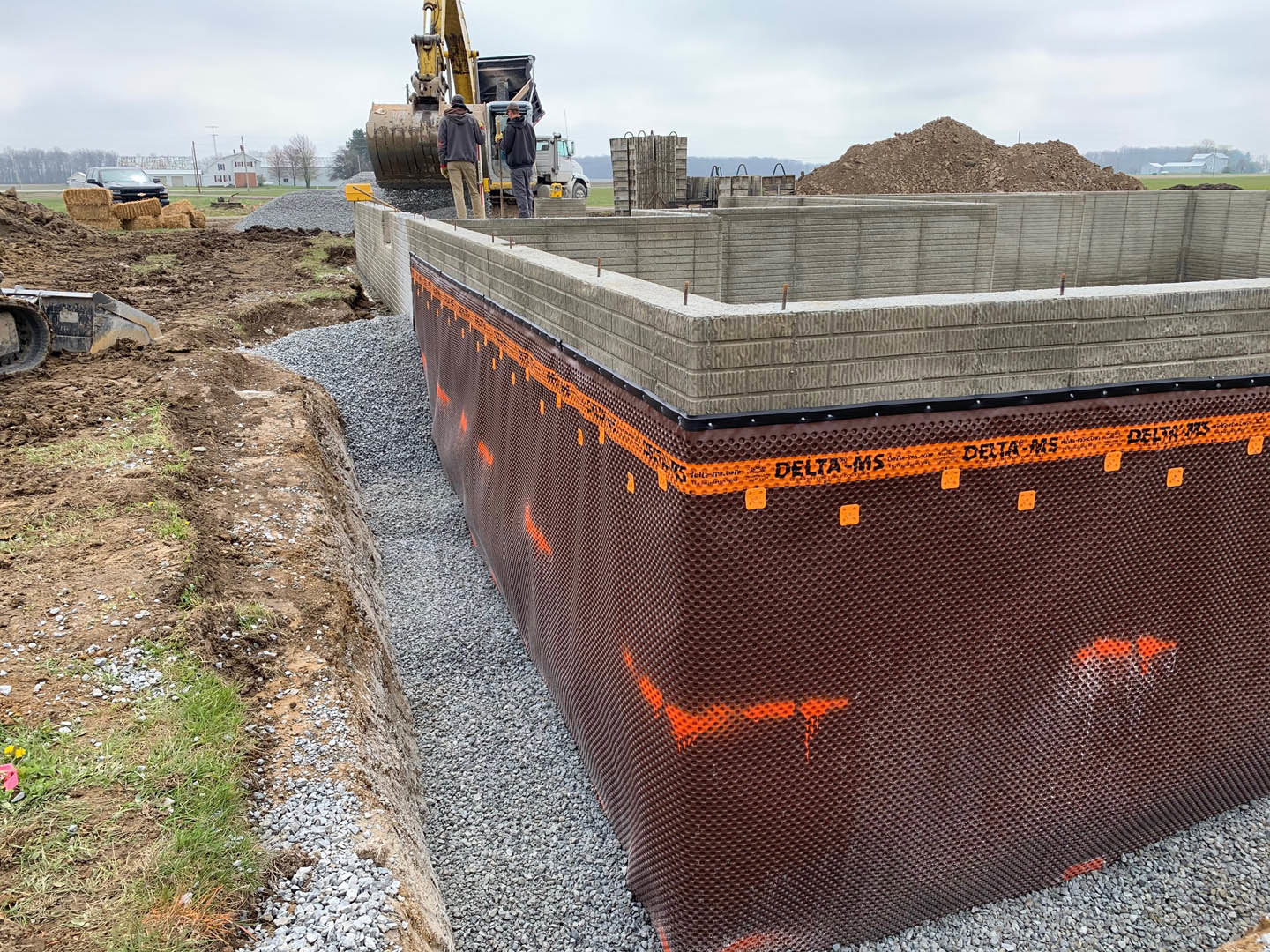 Concrete foundation and partially built wall on a residential construction site, large crane overhead, man in hoodie standing near wall, cloudy sky in background