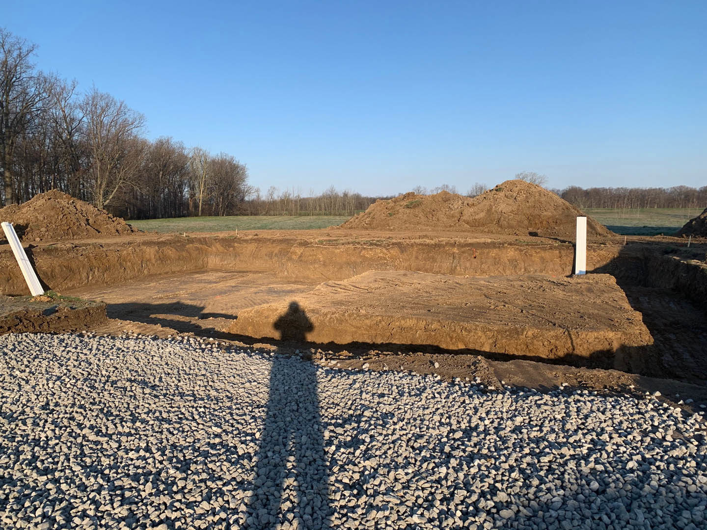 Person’s shadow cast across gravel and rocks, metal beam in foreground, leafless trees and blue sky with clouds in background
