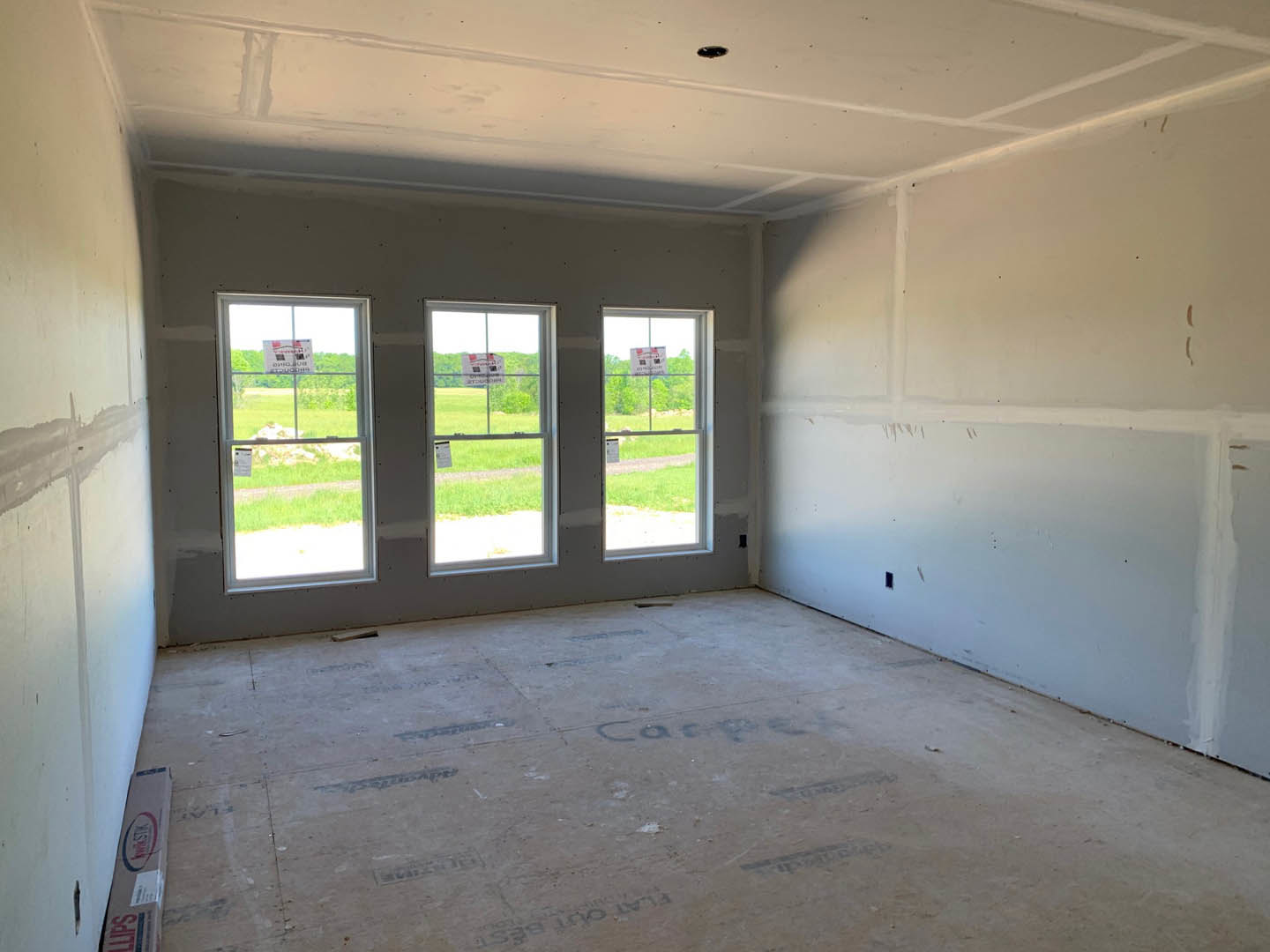 Bright room featuring a row of large windows with signs displayed, smooth plaster walls, light-colored flooring with words written on the surface, and a box positioned near a flag