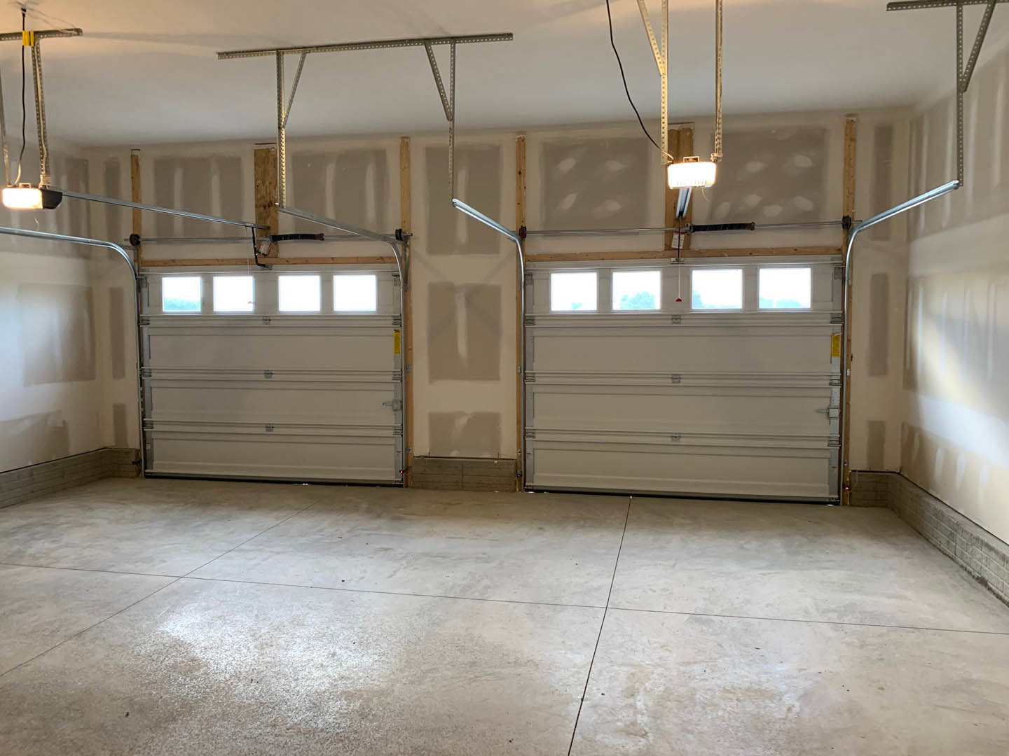 Spacious garage with two paneled garage doors featuring upper windows, concrete floor, white plaster walls, exposed ceiling beams, and a red ceiling fixture.
