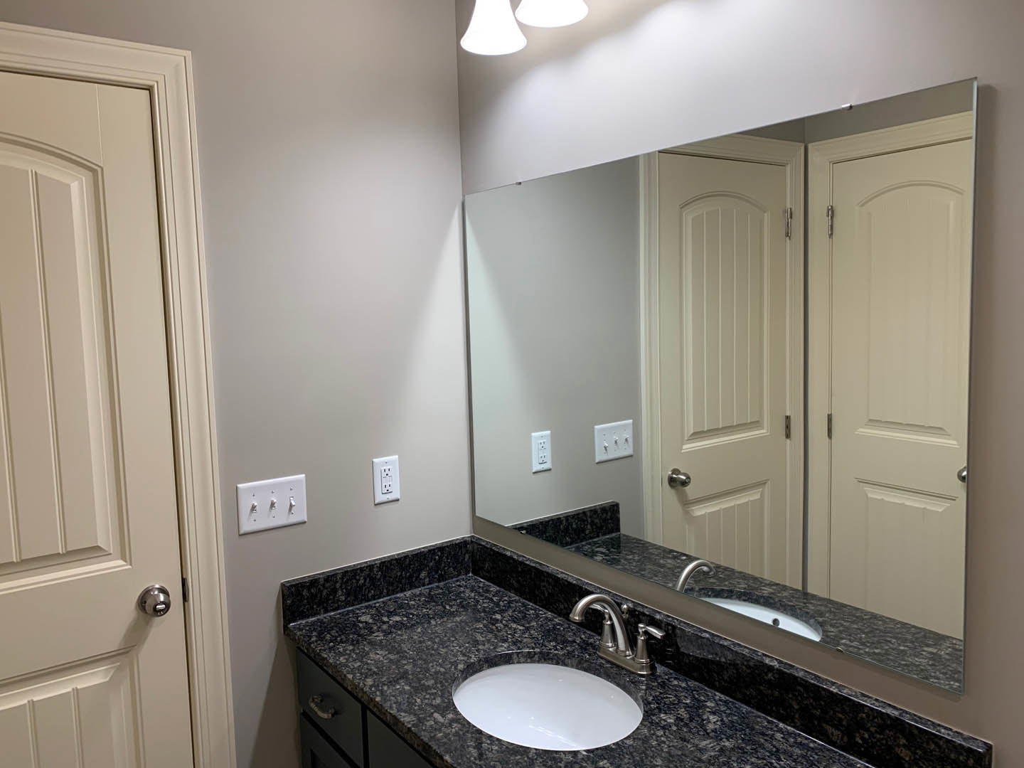 Modern bathroom featuring a rectangular mirror above a white sink with chrome faucet, black countertop, white tile walls, and a three-switch light panel adjacent to a white door
