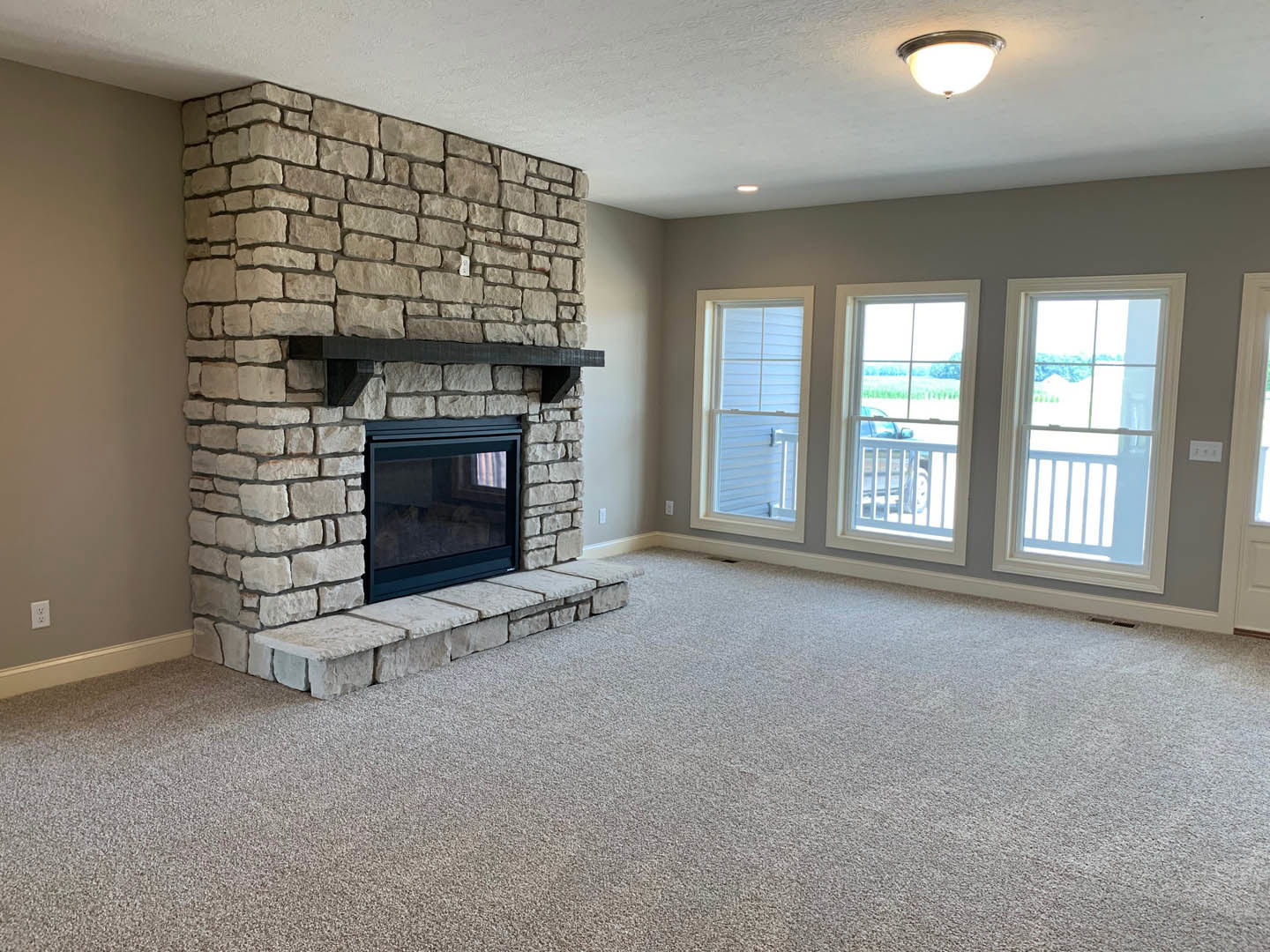 Carpeted living room featuring a wood-burning fireplace with glass doors and a wooden mantel, flanked by large windows with white trim and a railing