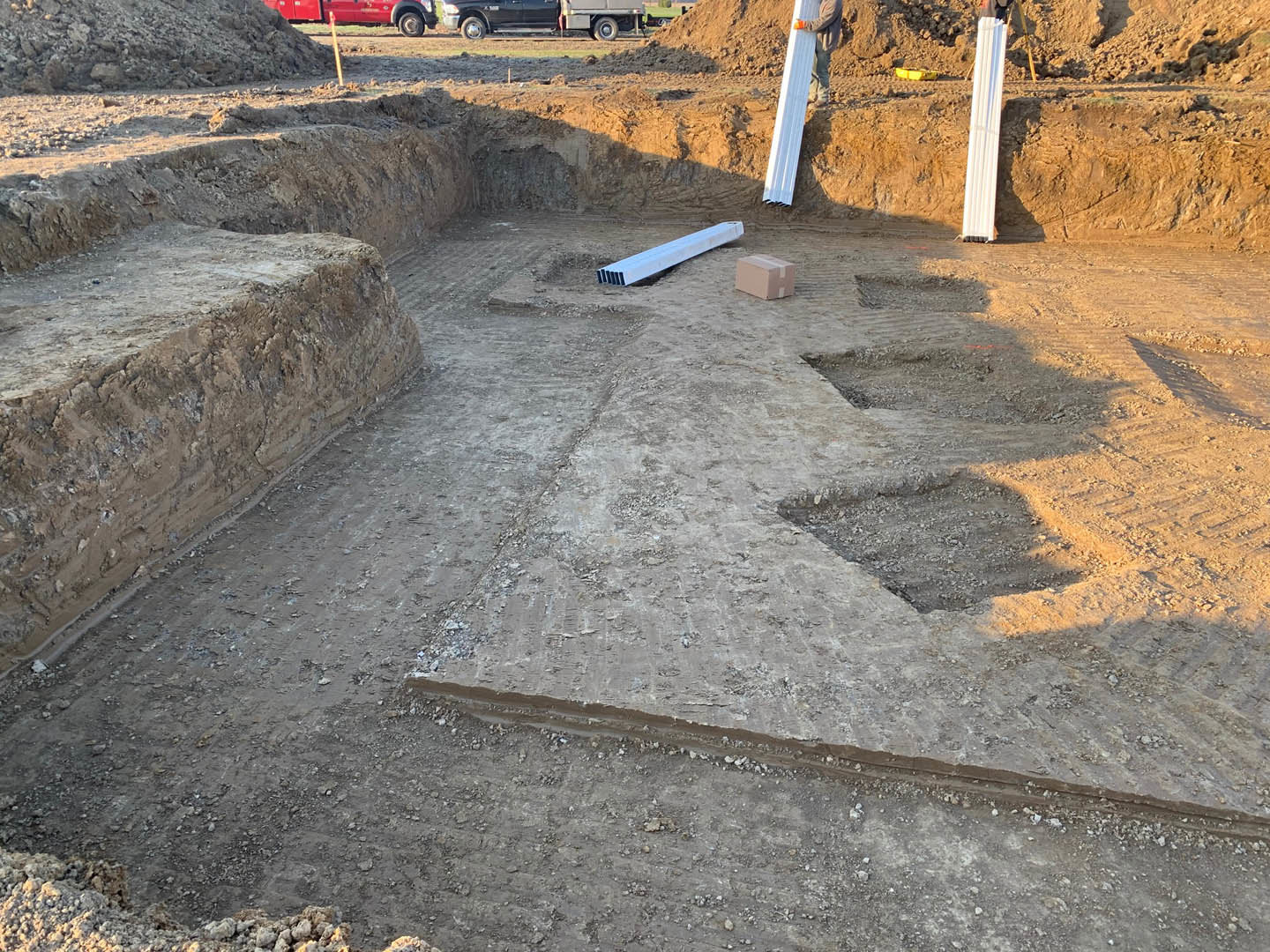 Construction site with exposed soil, scattered dirt piles, metal beams, concrete surfaces, and a white rectangular post on the ground