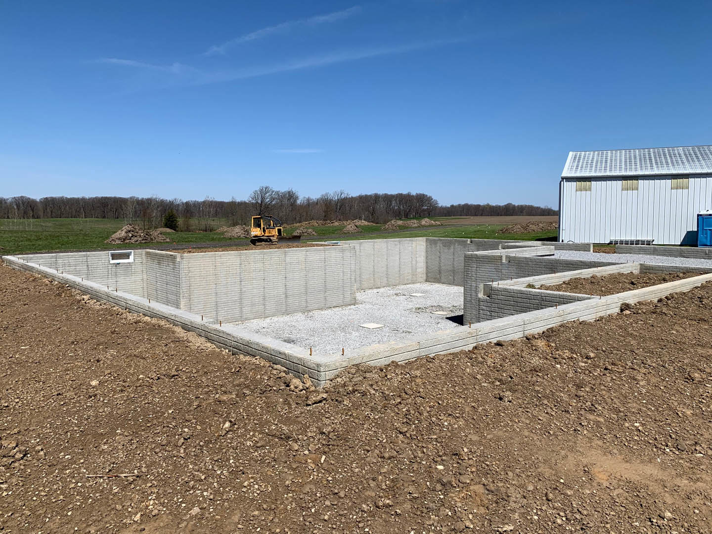 Concrete foundation and framing of a custom home under construction, gravel pile in foreground, yellow tractor parked near white barn with solar panels, blue sky and scattered