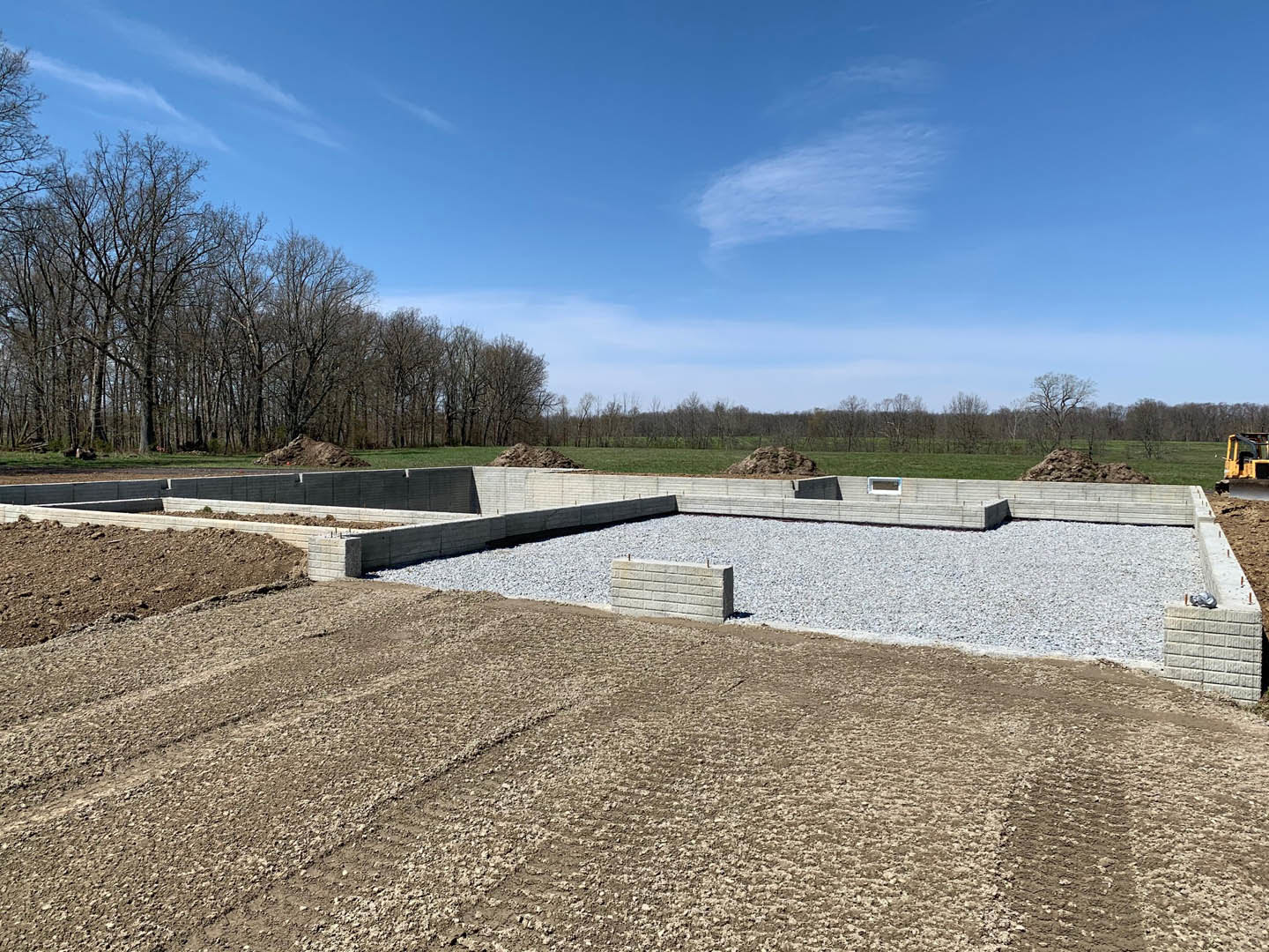 Concrete foundation surrounded by piles of gravel, white brick wall, scattered trees, and blue sky with clouds; yellow construction vehicle visible in foreground.