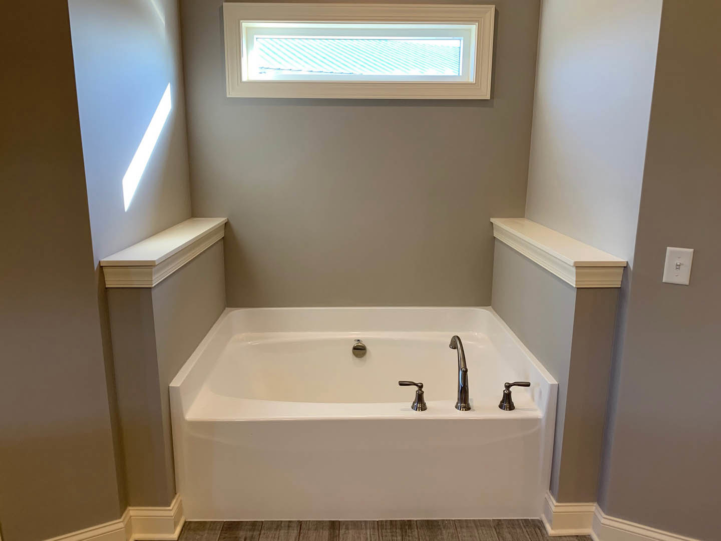 Freestanding white bathtub with chrome faucets and handle, positioned near a window with white trim, light switch on adjacent wall, black bell on white shelf above baseboard