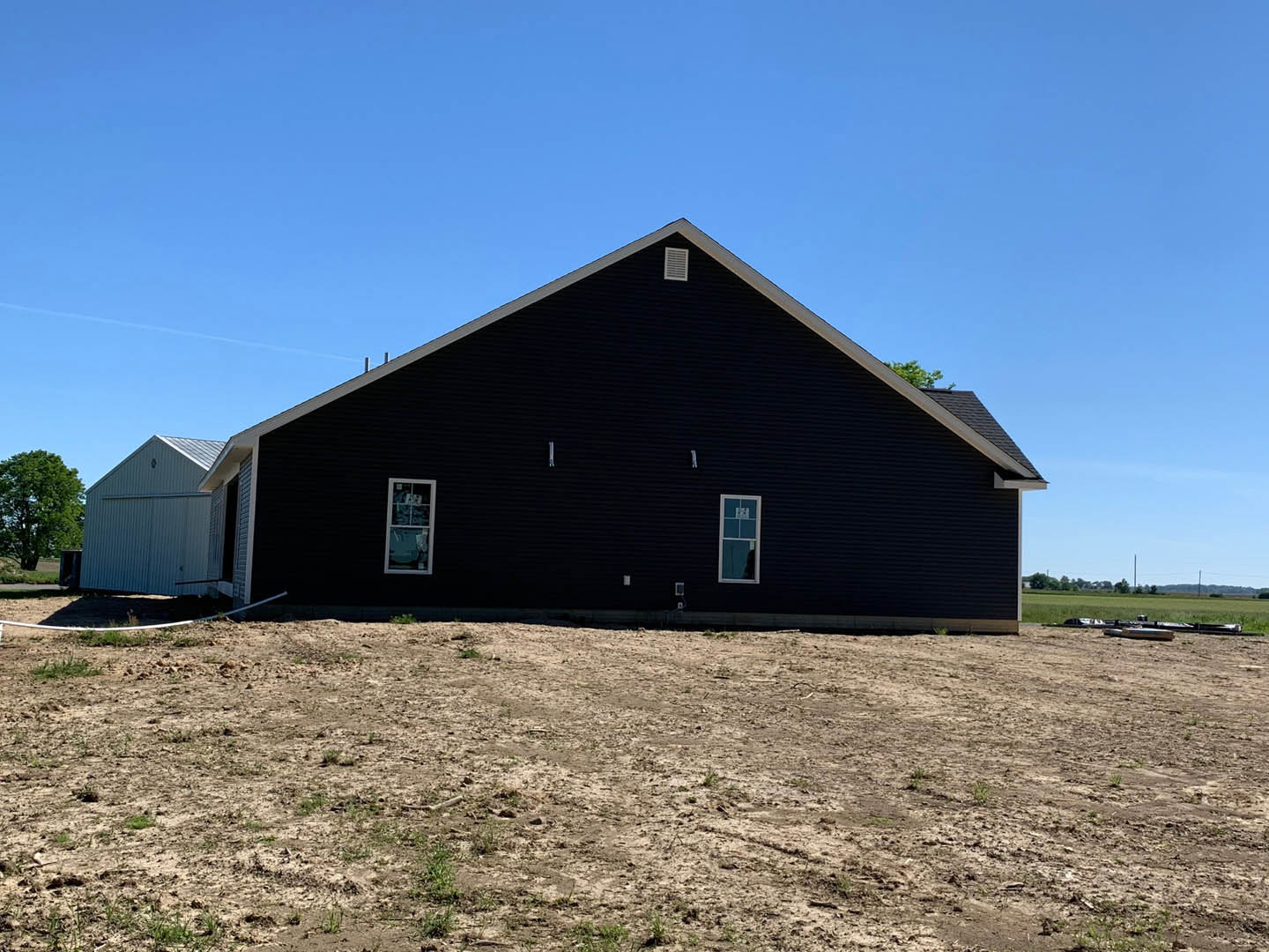Black house with white trim sits beside a dirt field with patches of grass, white-framed windows, nearby tree, and a white barn with a dark roof in the background.