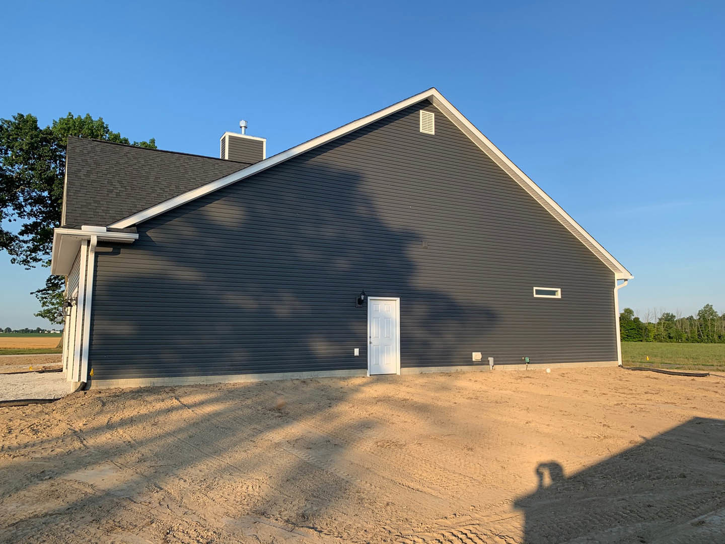 Single-story home with black siding, white front door, and dirt yard under clear blue sky; shadows of two people cast on ground, trees visible in background.