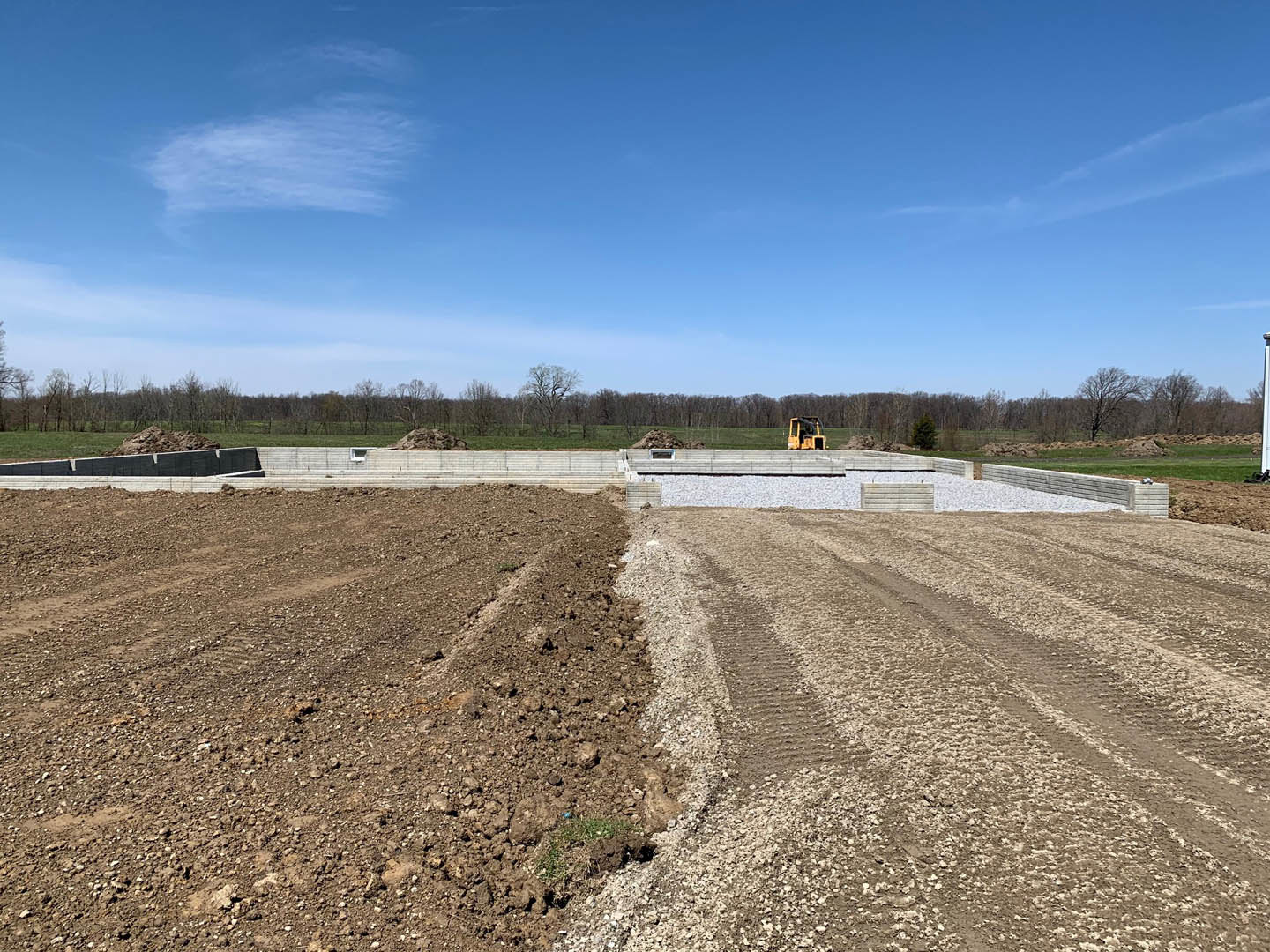 Dirt construction site with tire tracks, gravel, and a tractor under blue sky with scattered clouds, unfinished building in background
