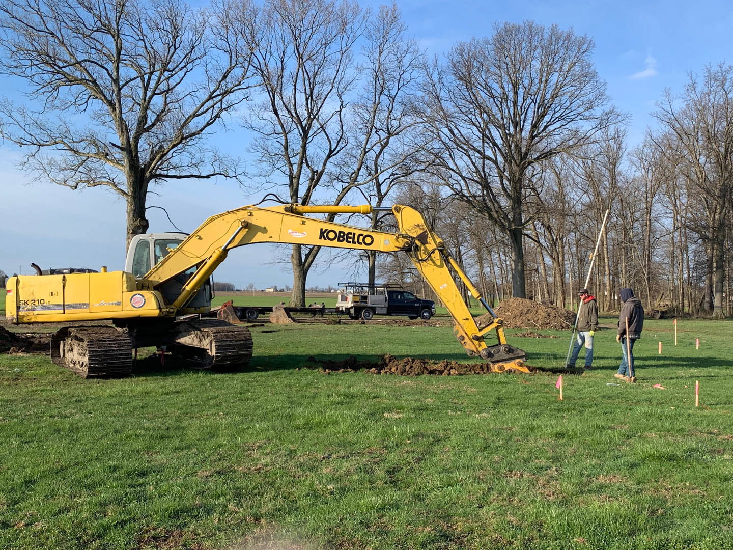 Yellow excavator with extended arm parked on grassy field near trees, construction worker standing beside machine holding shovel, blue sky overhead.