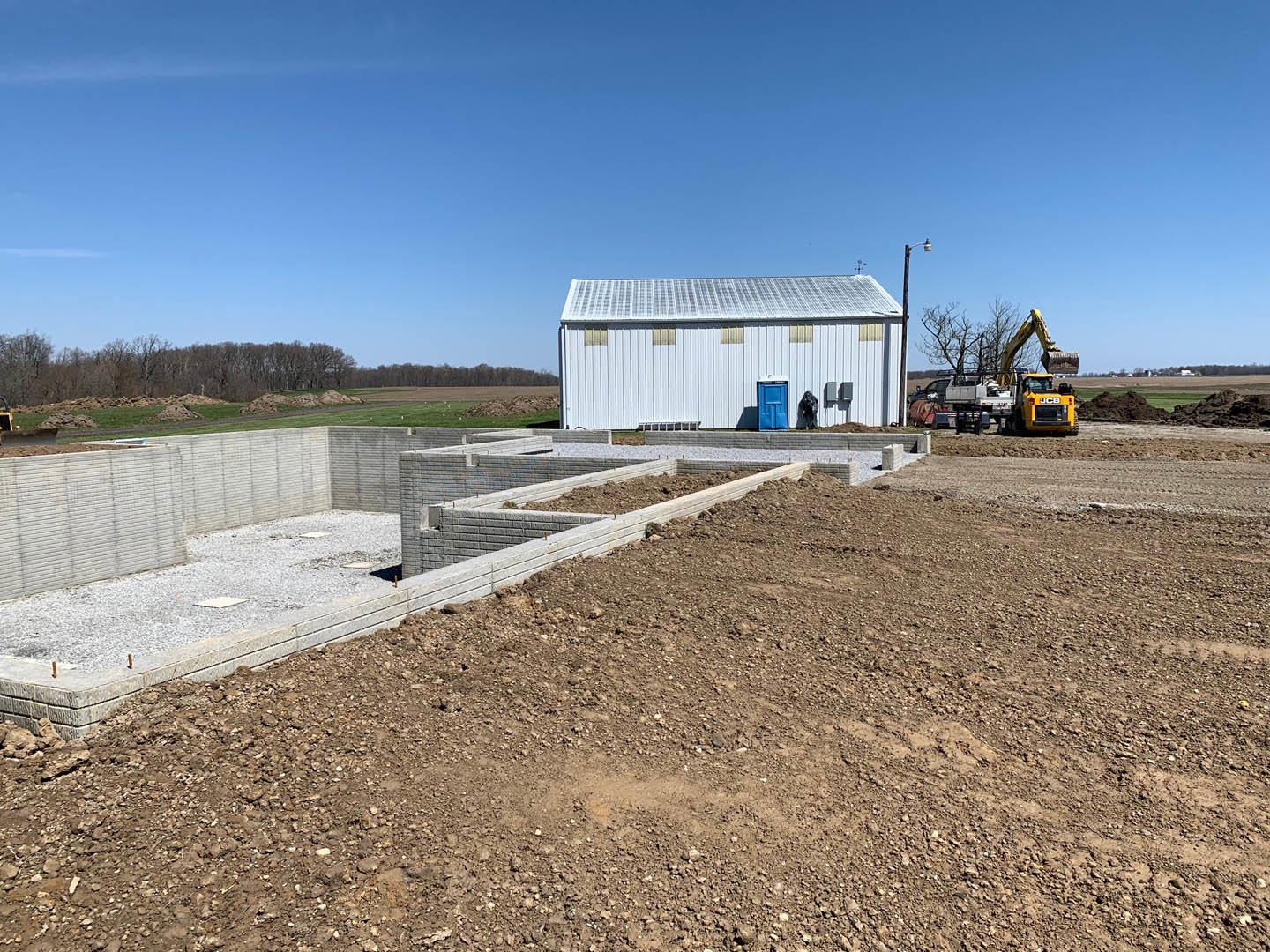White residential building under construction with exposed foundation, dirt patch in foreground, yellow construction vehicle, blue shipping container, and clear blue sky