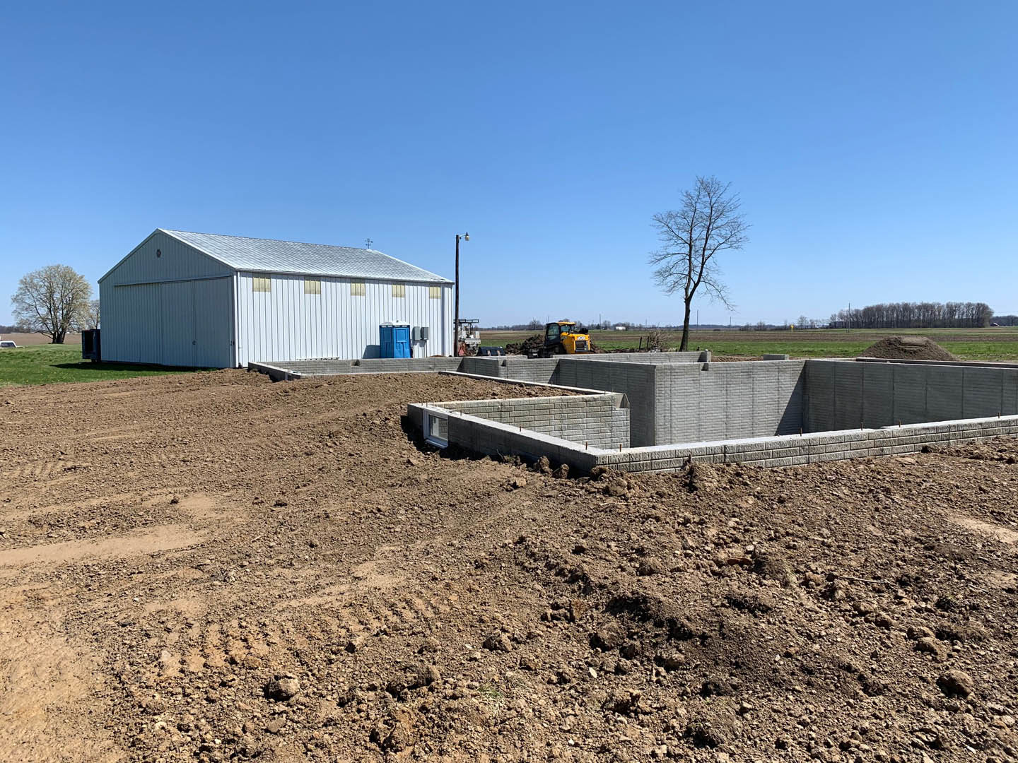 Modern white home under construction with exposed concrete walls, leafless tree, dirt lot, and tractor under clear blue sky