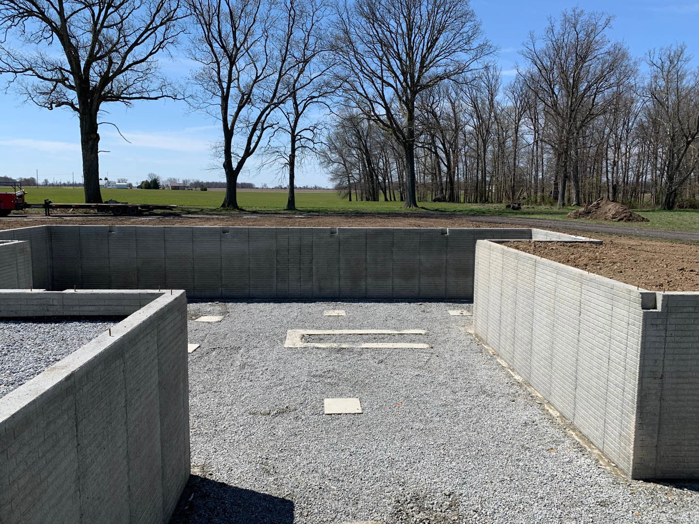 Concrete retaining wall with exposed aggregate finish, gravel pile at base, leafless trees and open field in background under clear sky