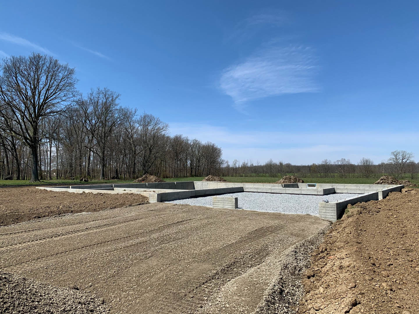 Concrete foundation with gravel base at a residential construction site, leafless tree and blue sky with scattered clouds in background