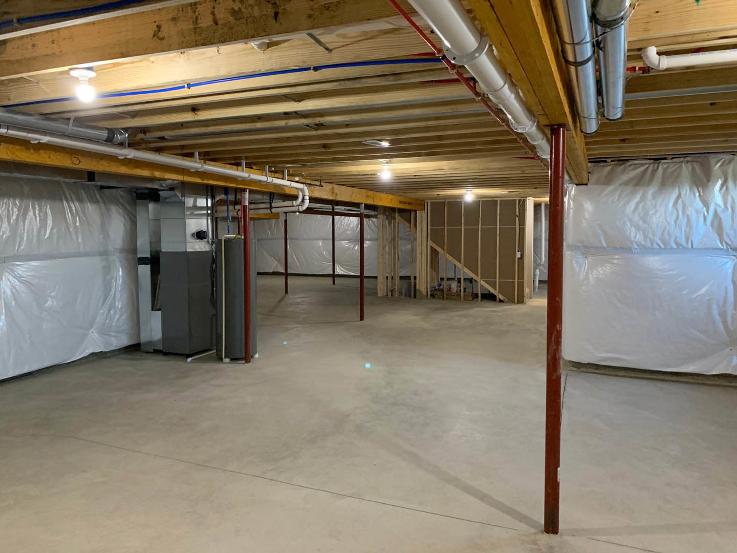 Unfinished basement with exposed ceiling pipes, metal utility box mounted on wall, wooden beam, white plastic bag on floor, and building insulation visible