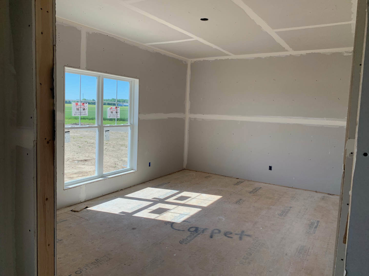 Bright room with two large windows, plaster walls, tile flooring, and sunlight casting a cross-shaped shadow on the concrete surface; window features signage with a house graphic
