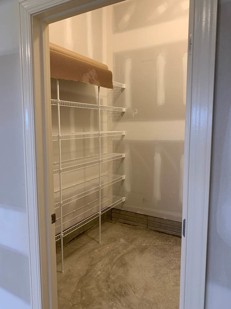 Walk-in closet with white shelves, concrete floor, white walls with black border, bed partially visible, window blinds in background