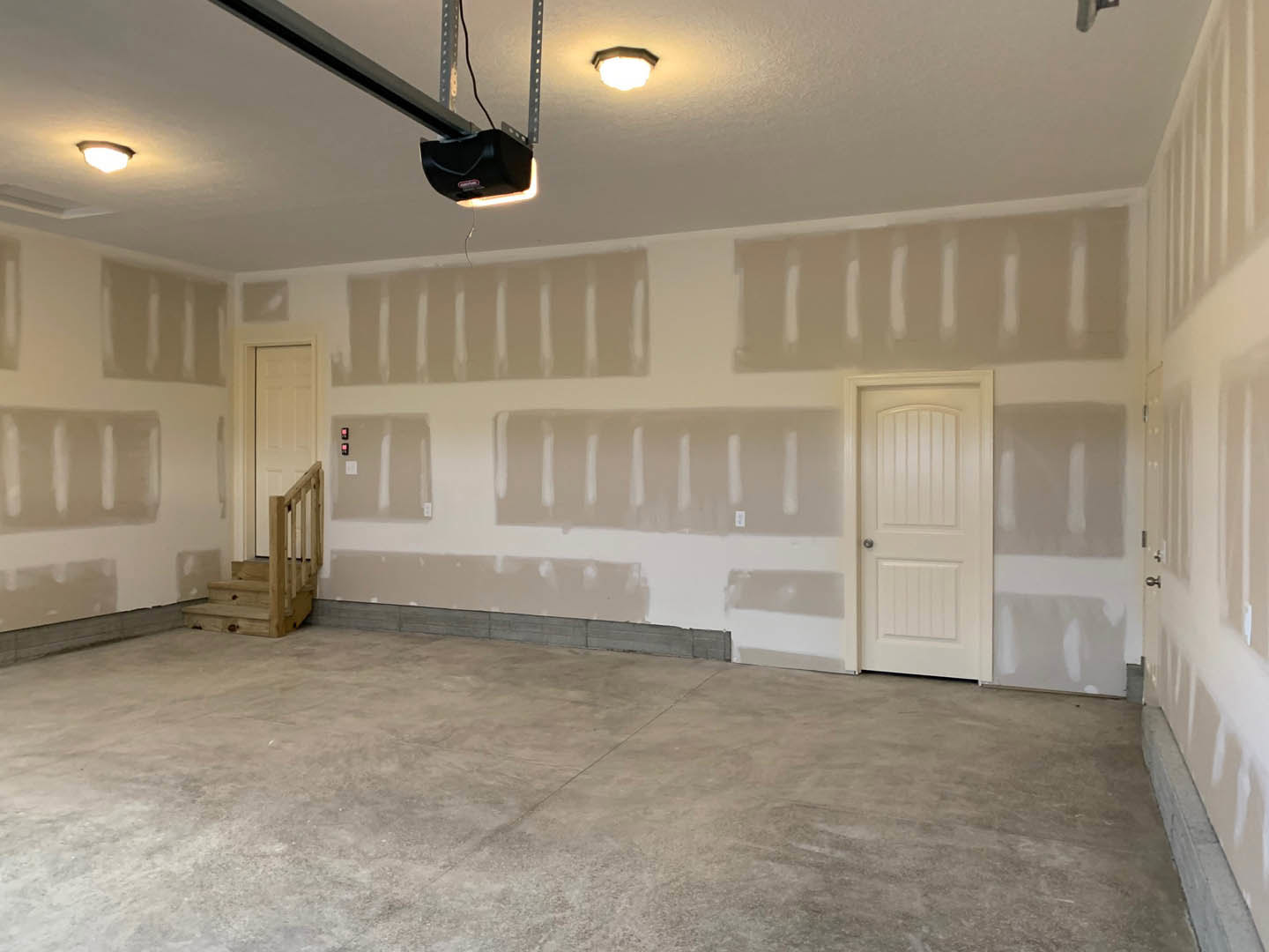 Concrete floor with a white door featuring a black handle, adjacent to a staircase; ceiling and wall lights, plaster walls, and a roller shutter visible.