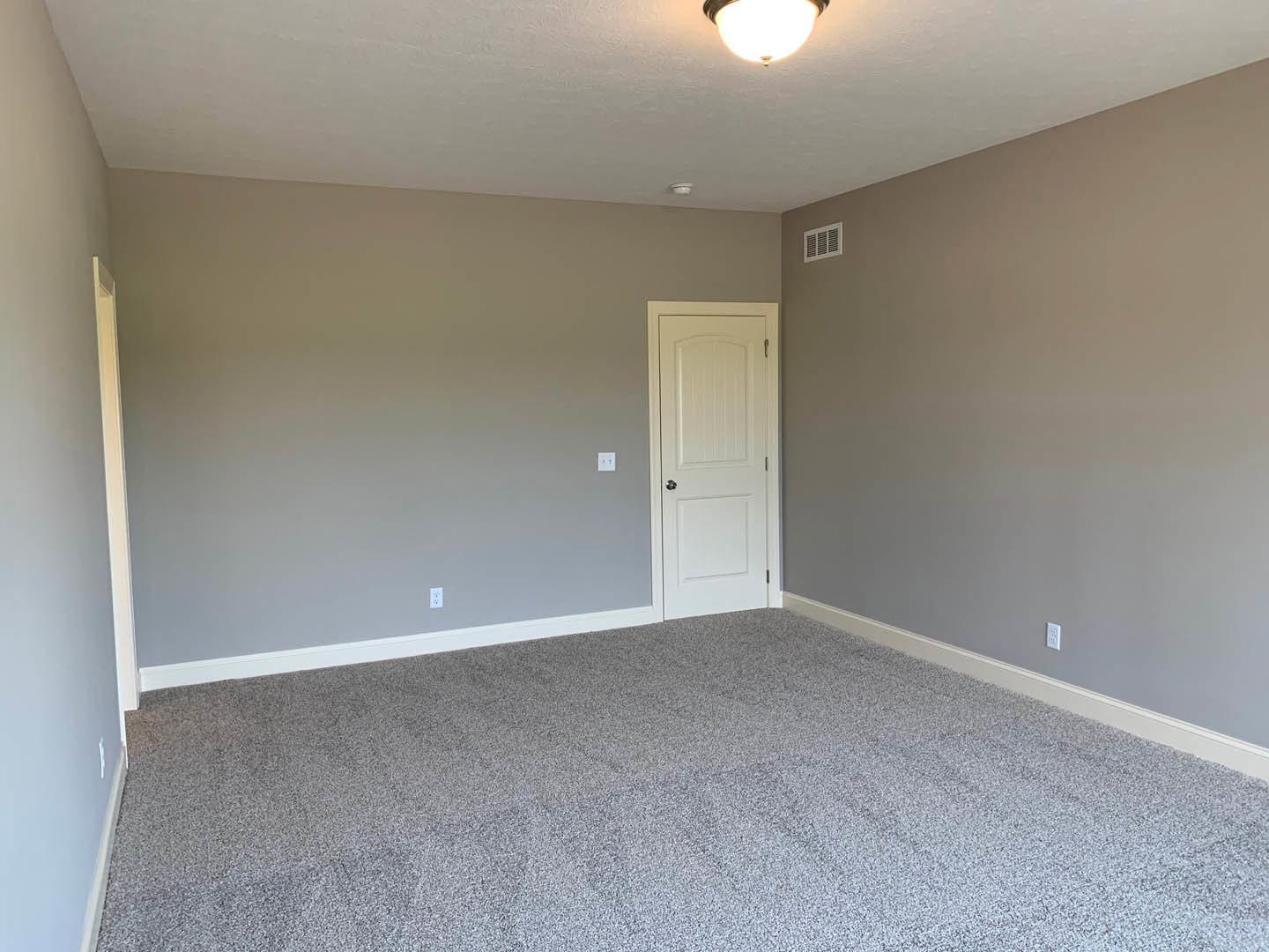 White paneled door with silver knob, beige carpeted floor, white walls with base molding, ceiling vent, flush mount ceiling light turned on