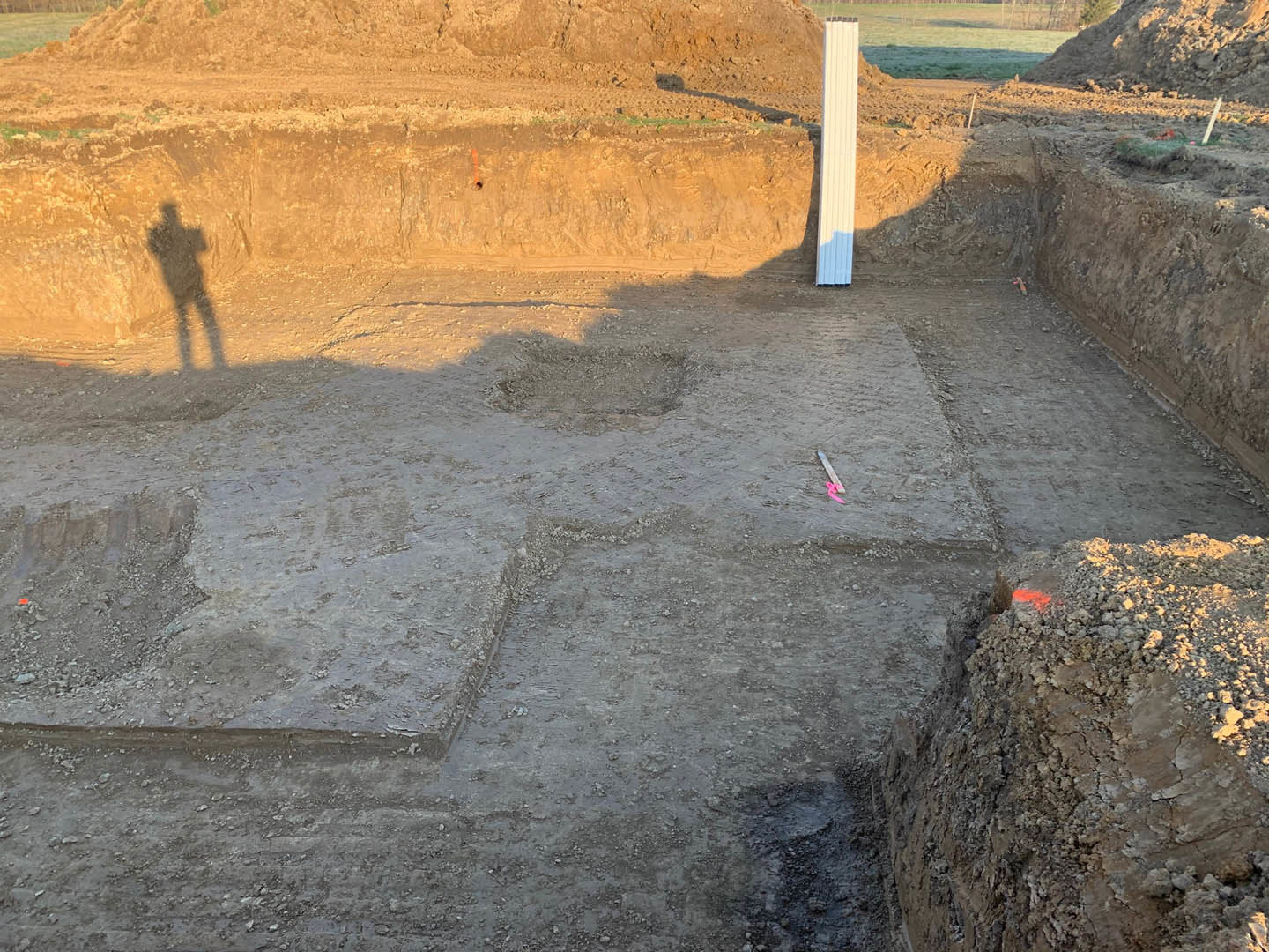 Shadow of a person cast on a dirt area near a large pile of soil, with a white rectangular object and a blue and white vertical window visible in the background.