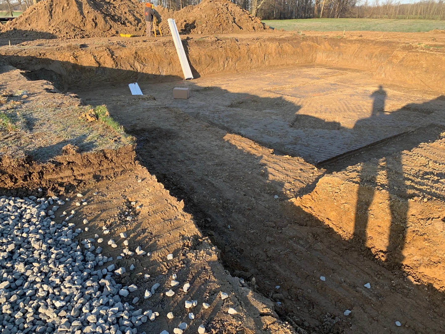 Construction site with exposed soil, scattered gravel piles, metal beams, and several workers in safety helmets standing near unfinished foundation.