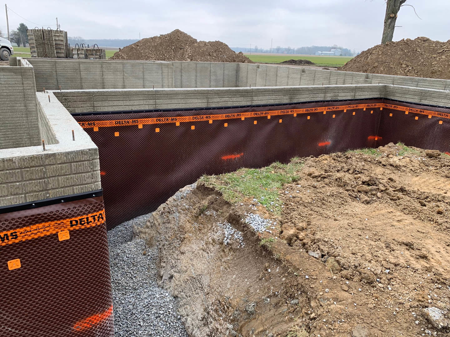 Brown and orange striped tarp covering a pile of dirt on a grassy construction site, fenced perimeter, man standing on hill, plastic wrap and composite materials visible, trees and
