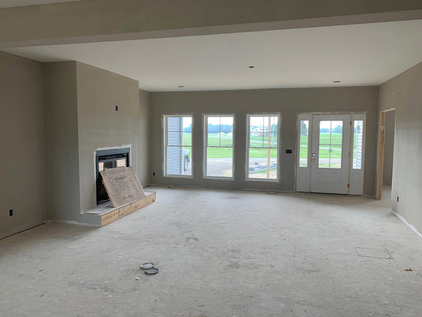 Living room with unfinished concrete floor, white fireplace, multiple windows, glass-paneled white door, and cardboard sign taped to window.