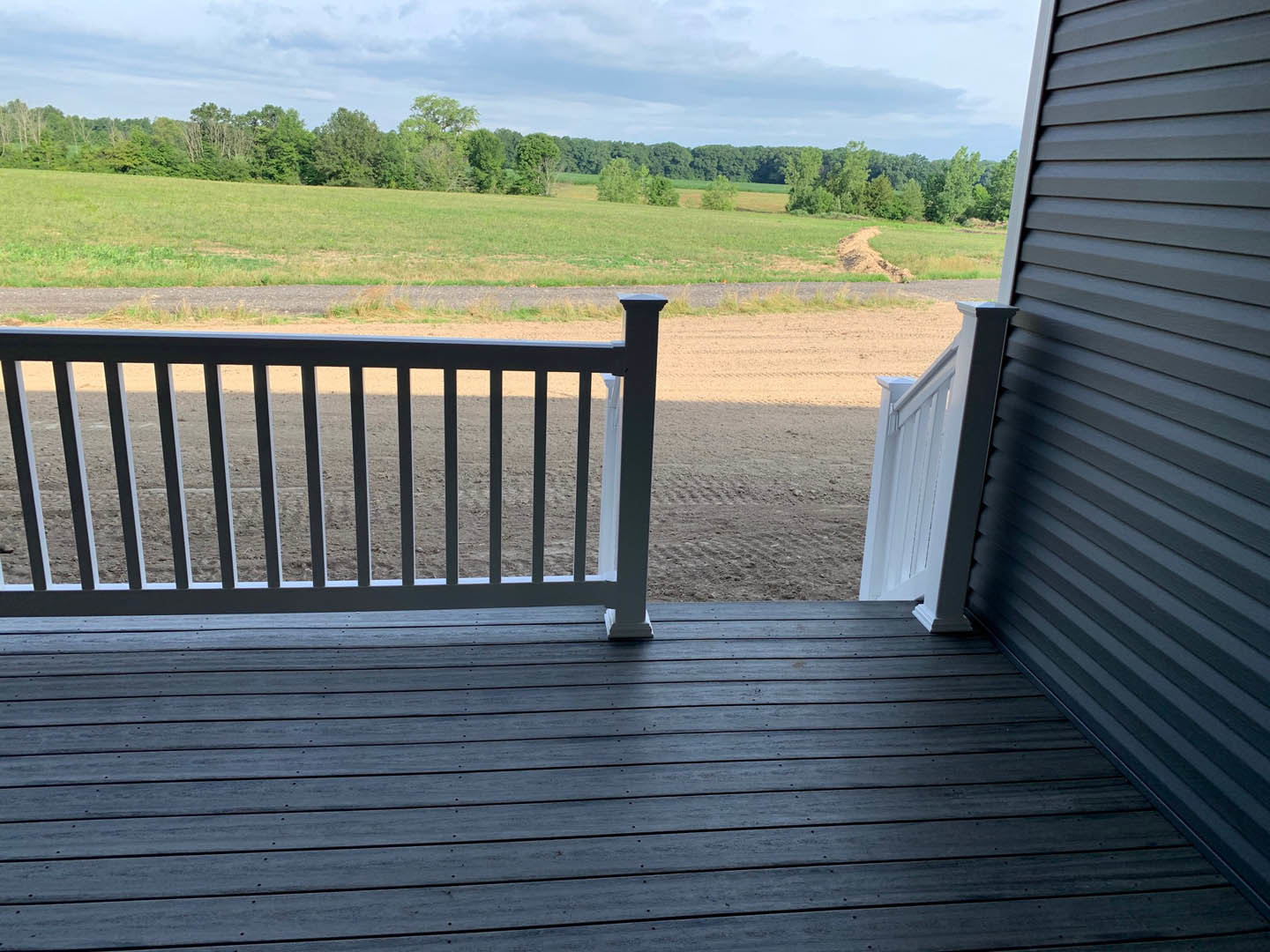 Wooden deck with white posts and railing overlooking a grassy field bordered by trees under a partly cloudy blue sky