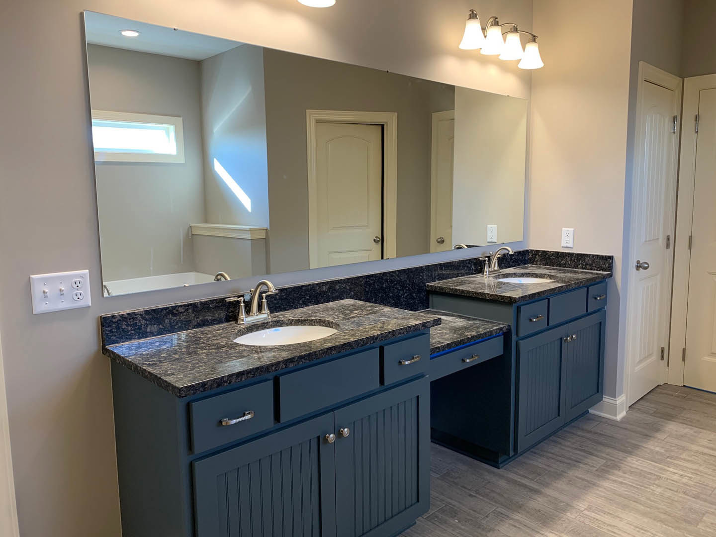 Bathroom with a wide mirror above a tiled countertop, white cabinetry and drawers, curved-top white door, modern light fixture, and a white outlet; person visible near a window