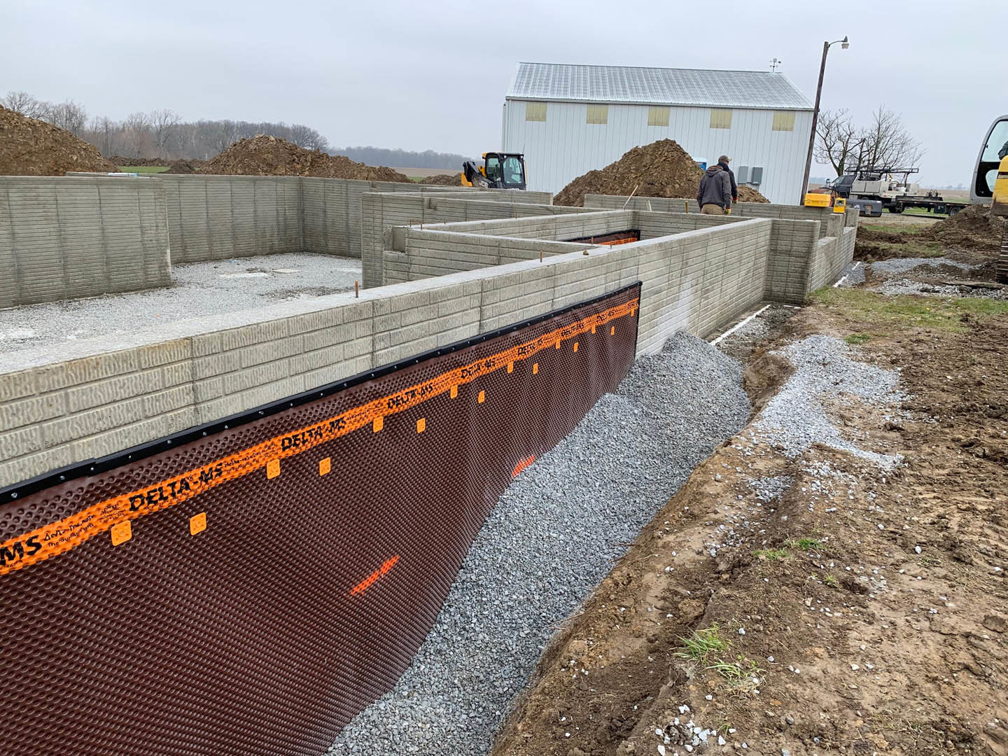 Brick exterior wall under construction with orange and black tape, pile of dirt in foreground, person in grey jacket near building with roof, construction vehicle close by, trees
