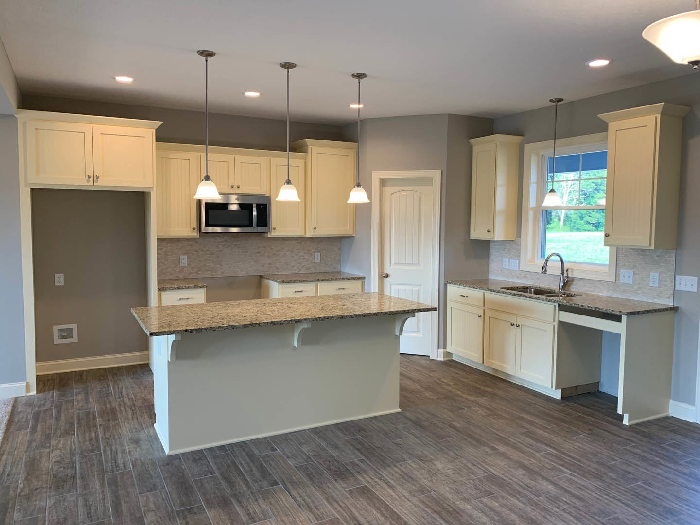Spacious kitchen featuring a large central island with white cabinetry, stainless steel microwave, wood flooring, white walls, and a white door with a silver knob.