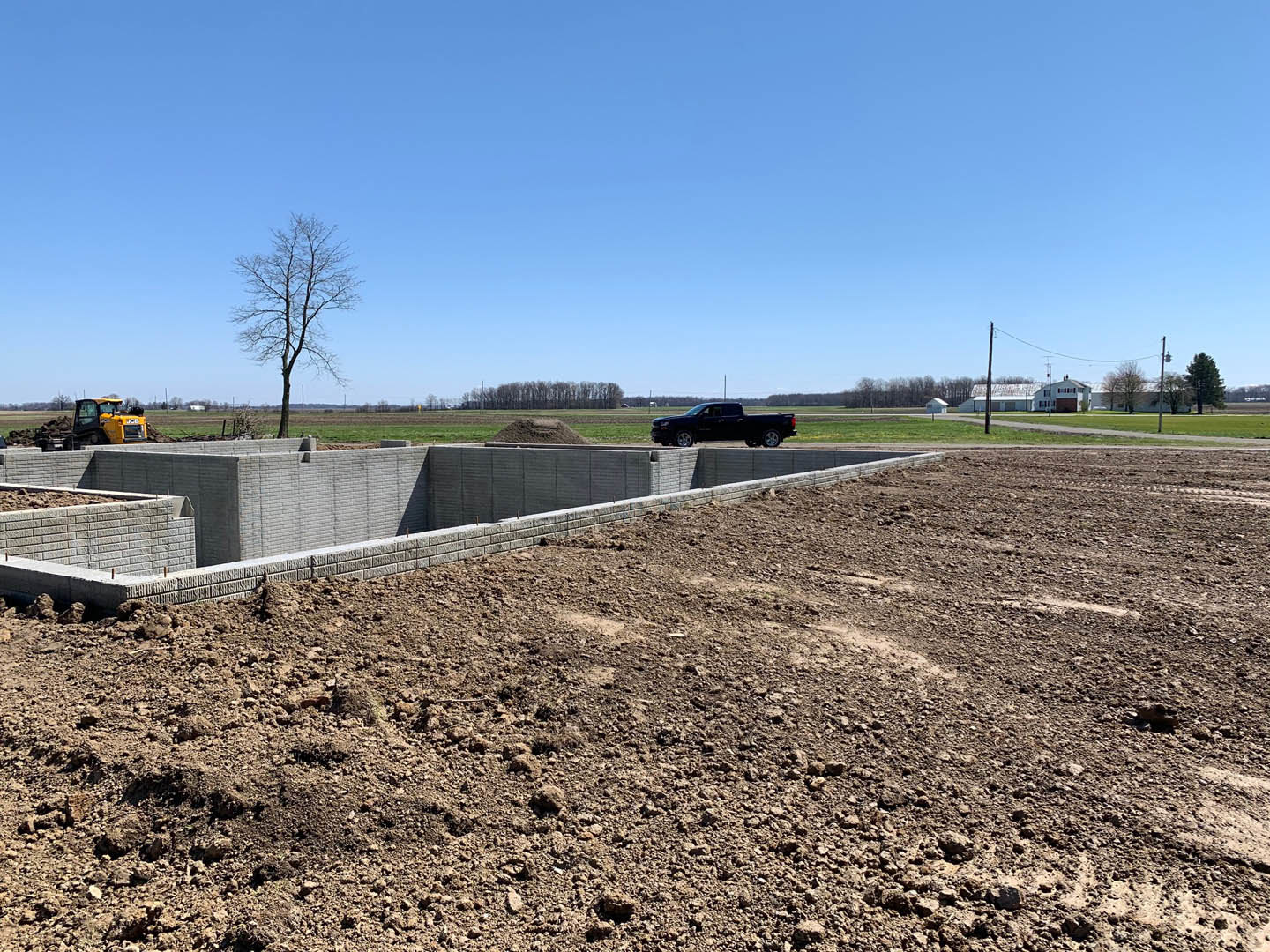 Black pickup truck driving through excavated hole in dirt, leafless tree nearby, concrete wall and grassy field under clear blue sky