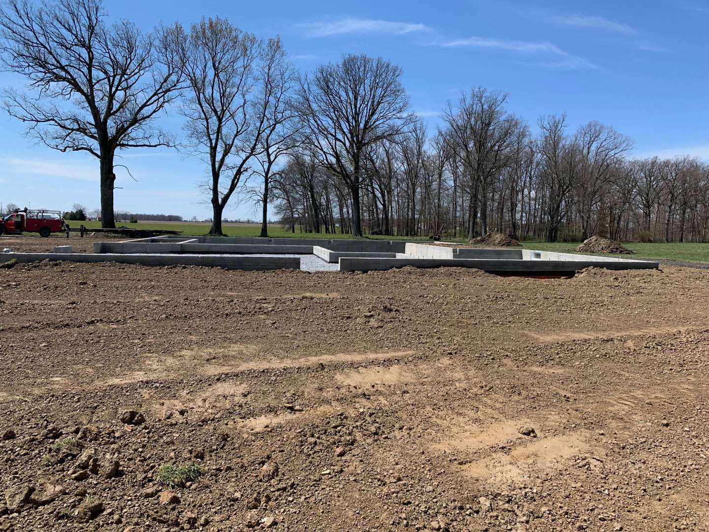Dirt field bordered by leafless trees, red truck with ladder parked near scattered concrete blocks, gravel and grass patches along a low concrete block wall