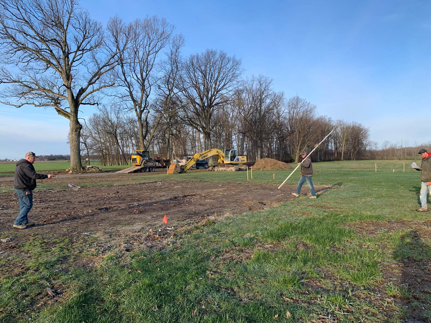 Modern home construction site with grassy field, exposed soil, scattered trees, and a man in a black shirt holding a long pole near construction equipment