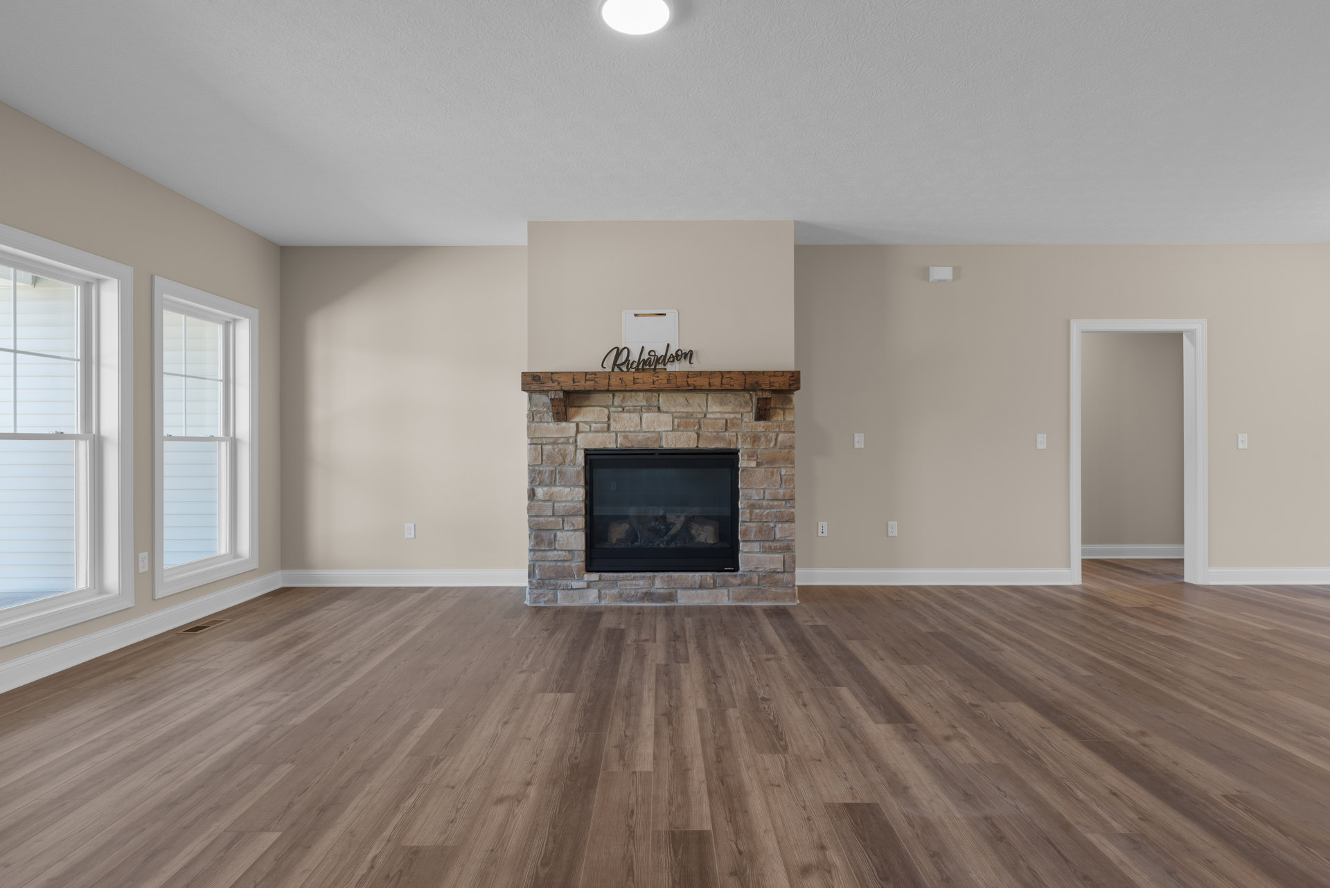 Living room with hardwood floors, white-framed fireplace featuring a wood beam mantel and stacked logs, grey accent wall, window with blinds