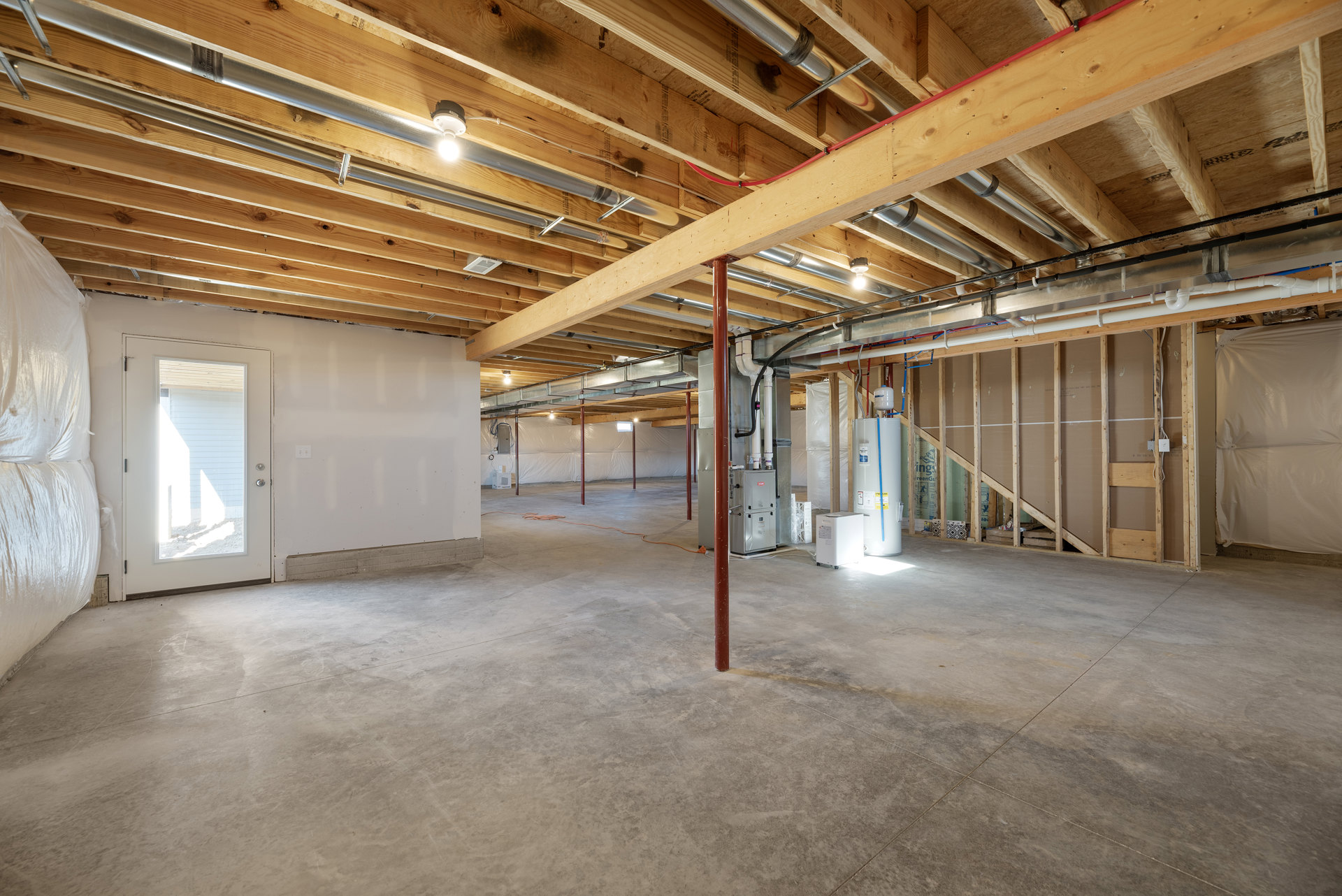 Room with exposed wood ceiling, concrete floor, door with window, white box on wheels, red poles, and white plastic sheet covering part of wall.
