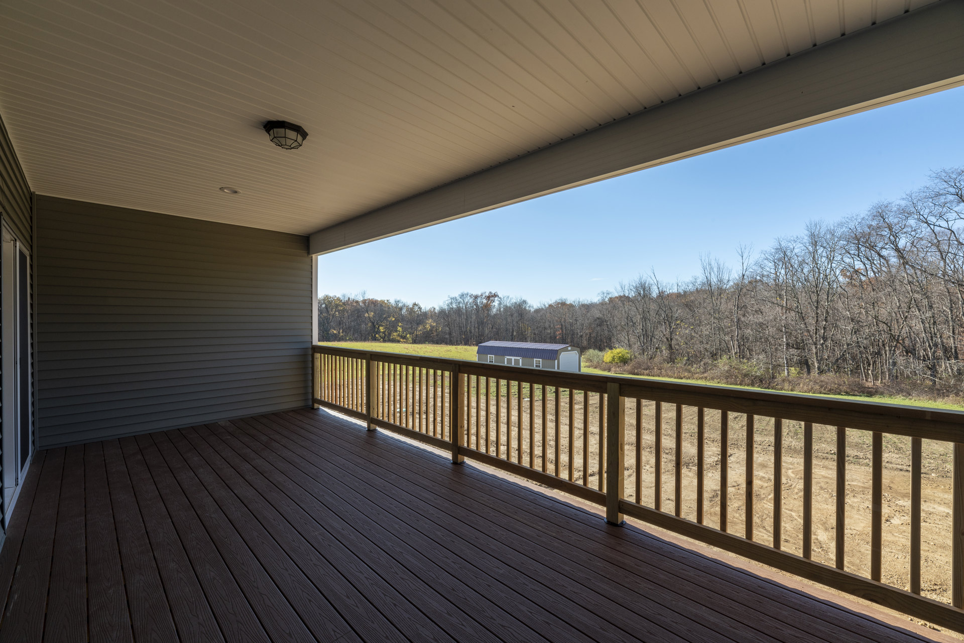 Wood deck with composite railing overlooking a field of mature trees, blue metal roof visible, outdoor light fixture mounted near entry.