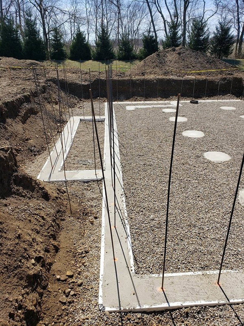 Gravel and concrete foundation with circular markings, several dirt piles, surrounded by trees and soil under a clear sky