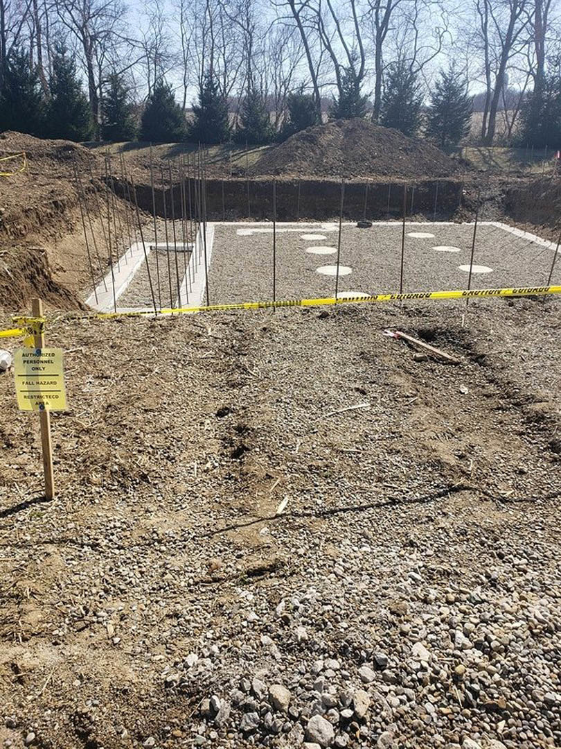 Construction site with fenced perimeter, yellow caution tape on poles, row of white markers, dirt pile on hill, yellow warning sign, and surrounding forested trees.