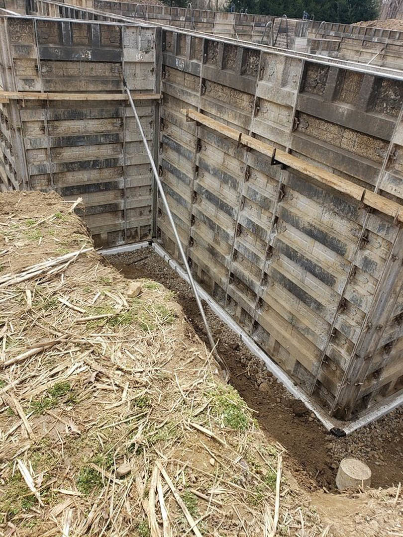 Wooden wall with vertical metal pole, grassy ground, straw pile, and partial view of wooden beams and ladder at construction site.