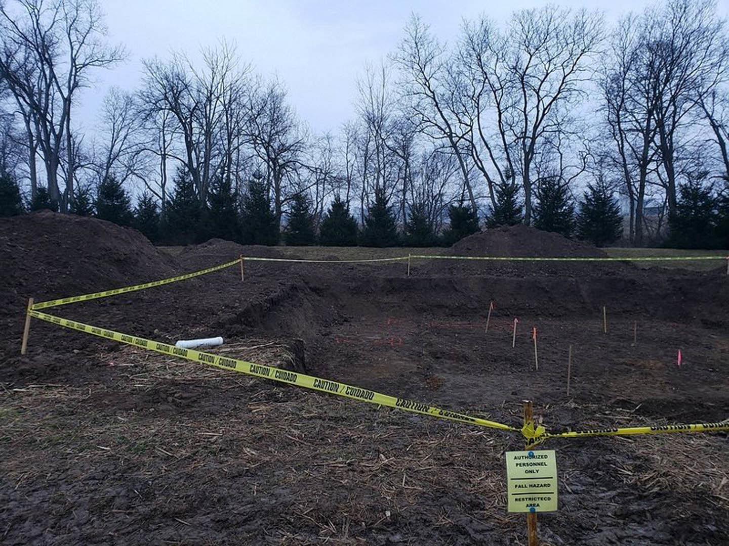 Dirt pit surrounded by yellow caution tape on a residential construction site, bare trees in background, yellow warning sign visible, grassy ground and soil.