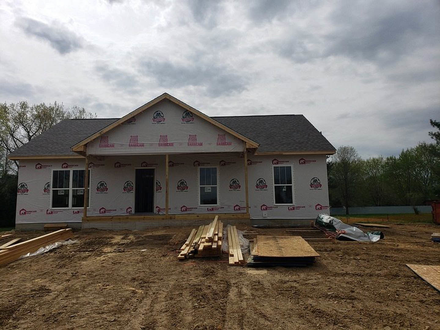 Wood-framed house under construction with black door, white trim, window displaying sign, person holding paper, and pile of lumber on ground