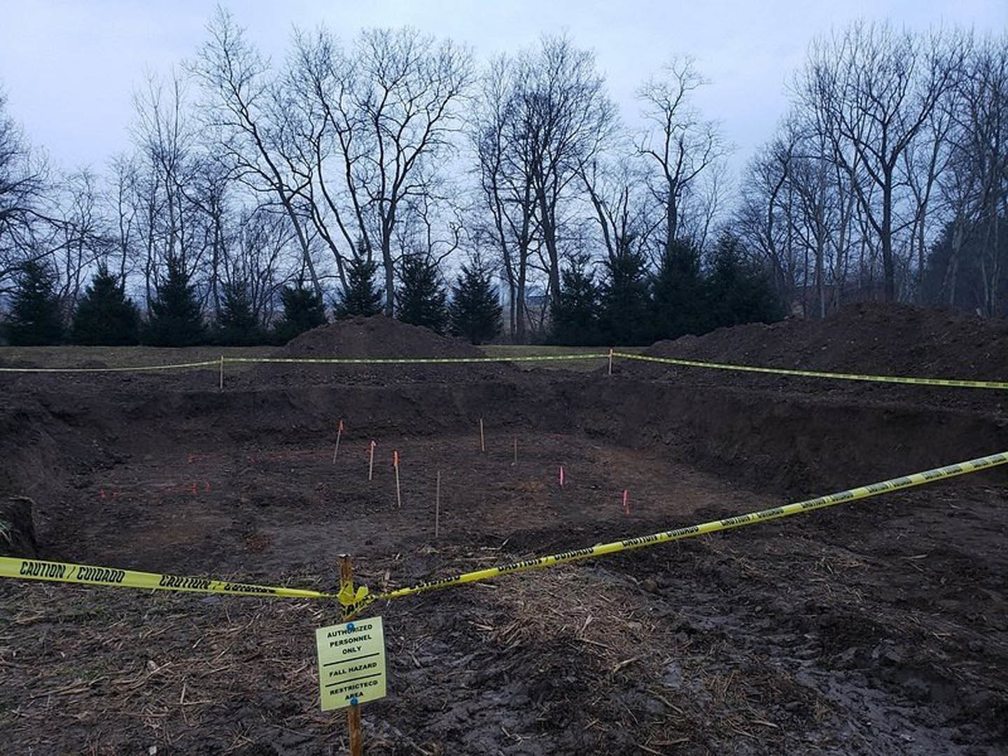 Dirt excavation surrounded by yellow caution tape, bordered by trees and grassy landscape, wooden post marking construction site boundary