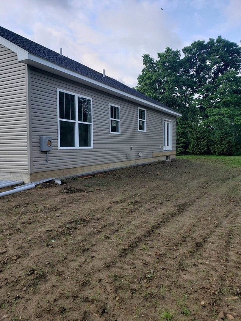 Two-story home with gray siding, several windows, surrounded by dirt lot and scattered trees, Plattsburgh Air Force Base visible in distant background under cloudy sky.