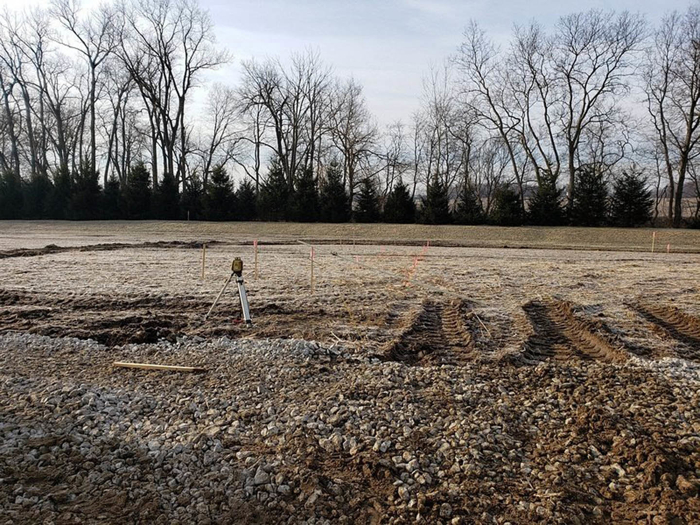 Dirt field with tire tracks, scattered clouds in the sky, row of trees lining the horizon, winter landscape with bare ground and distant fence