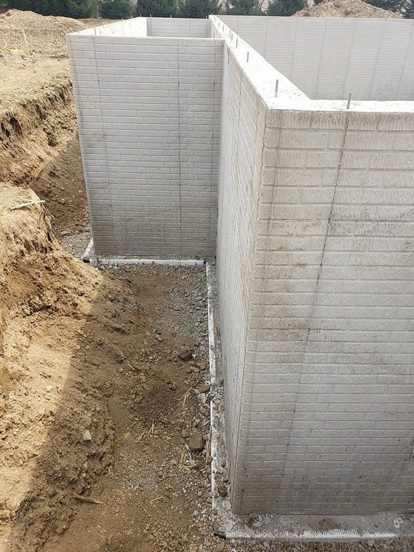 Concrete foundation wall with a large hole in the ground, surrounded by soil, rocks, and a pile of dirt.