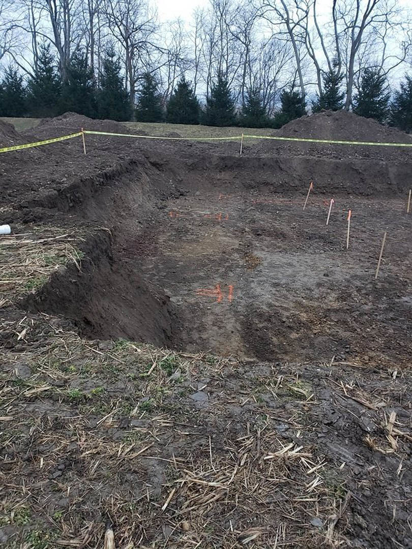 Excavated dirt pit surrounded by trees, yellow caution tape marking perimeter, clear sky overhead