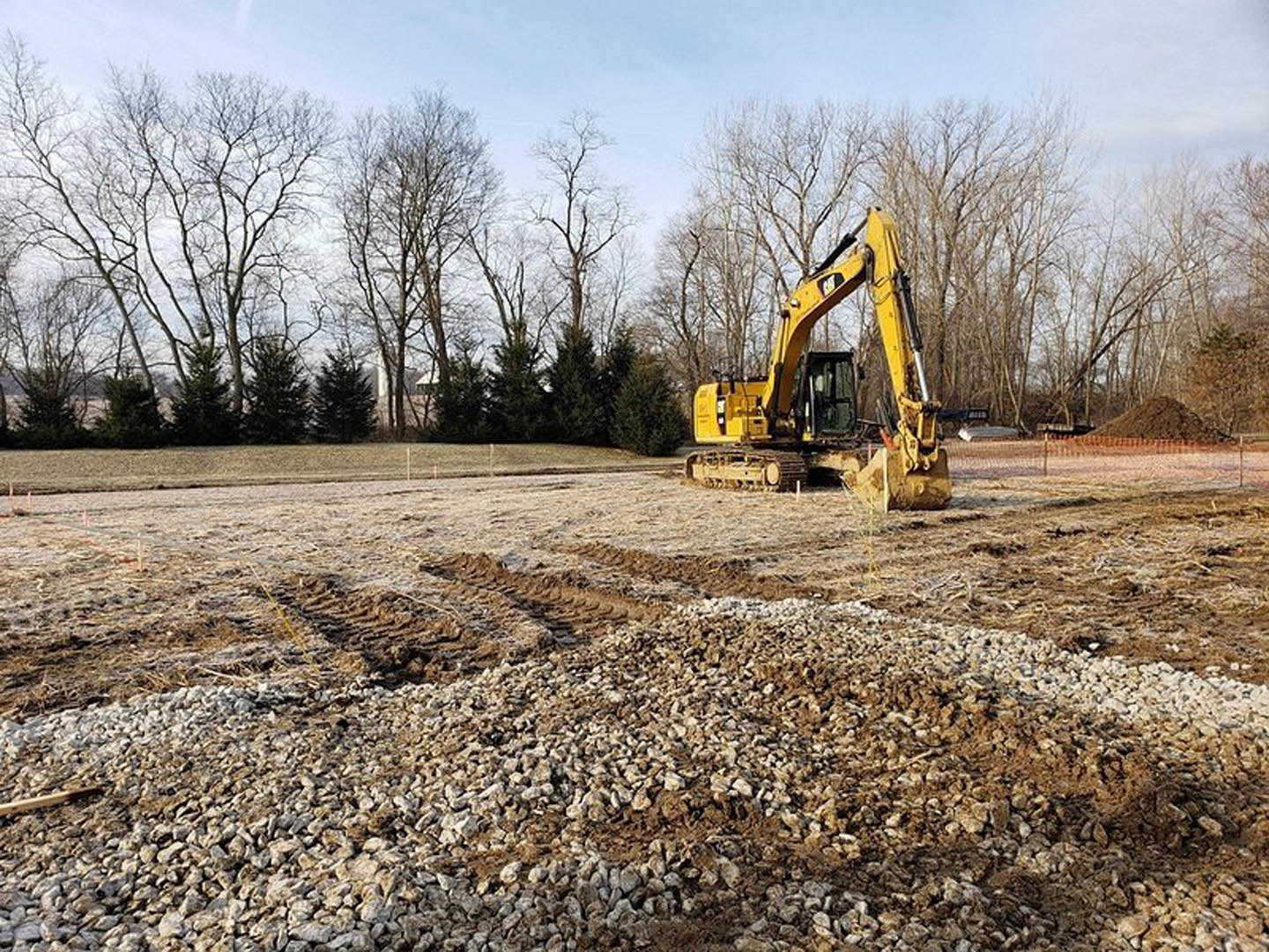 Yellow excavator parked on a dirt lot surrounded by trees, with piles of rocks and soil in the foreground
